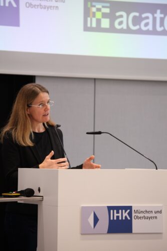 Anna Frey standing at the podium, welcoming the participants. A presentation can be seen on a screen in the background