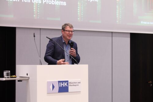 Hubert Jäger holding a microphone in his hand and standing in front of a screen on which his presentation can be seen
