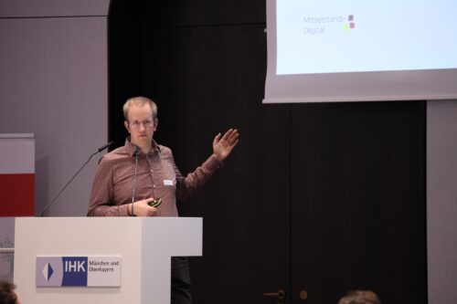 Dennis Huschenhöfer pointing at the presentation, which can be seen behind him, during his keynote